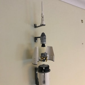 May include: A white and gray Lego model of a rocket ship, disassembled and mounted on a wall. The rocket is divided into four sections: the top, the command module, the service module, and the main engine. The model is mounted on a wall with black brackets.