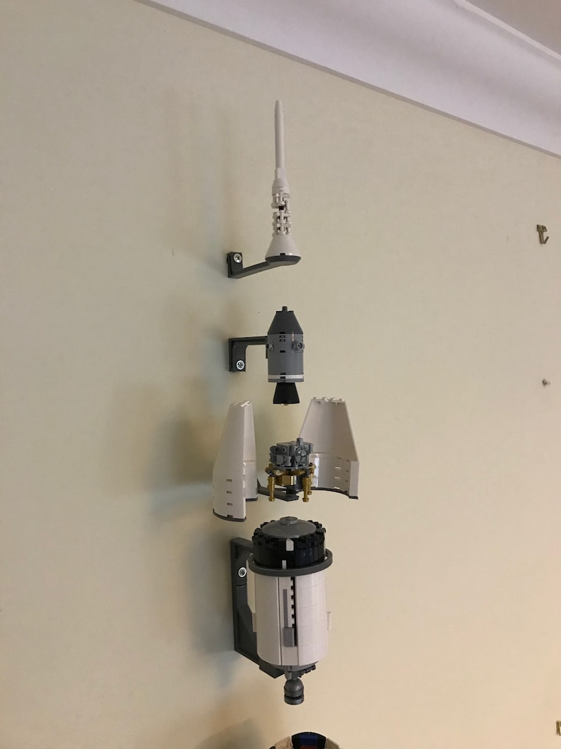 May include: A white and gray Lego model of a rocket ship, disassembled and mounted on a wall. The rocket is divided into four sections: the top, the command module, the service module, and the main engine. The model is mounted on a wall with black brackets.