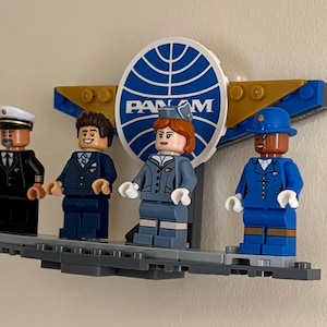 May include: A Lego display featuring four minifigures representing Pan Am airline personnel. The figures include a pilot, two flight attendants, and a porter, all standing in front of a Pan Am logo. The figures are on a gray platform.