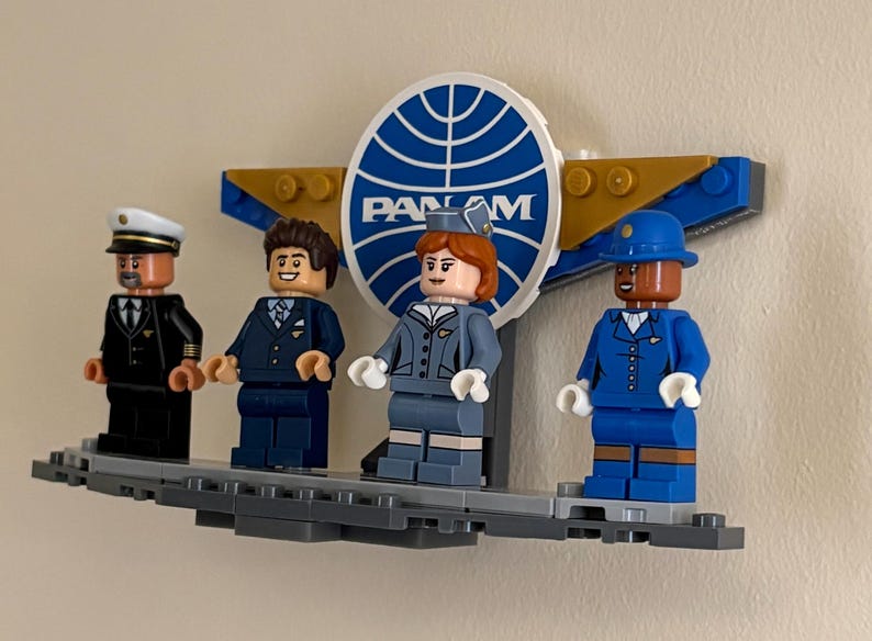 May include: A Lego display featuring four minifigures representing Pan Am airline personnel. The figures include a pilot, two flight attendants, and a porter, all standing in front of a Pan Am logo. The figures are on a gray platform.
