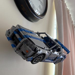 May include: A silver and blue toy car model with detailed features, including wheels and a visible engine. The model is constructed from interlocking blocks, showcasing a sleek design. The car is displayed against a white wall, with a clock in the background.