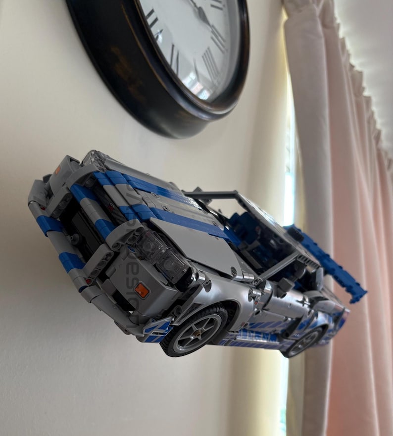 May include: A silver and blue toy car model with detailed features, including wheels and a visible engine. The model is constructed from interlocking blocks, showcasing a sleek design. The car is displayed against a white wall, with a clock in the background.