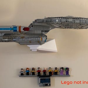Puede incluir: Un modelo gris de una nave espacial, montado sobre un soporte blanco, con una peque&ntilde;a estanter&iacute;a debajo que contiene figuras en miniatura. El modelo tiene detalles en azul y naranja. La estanter&iacute;a tambi&eacute;n tiene una peque&ntilde;a placa. El texto "Lego not included" es visible.