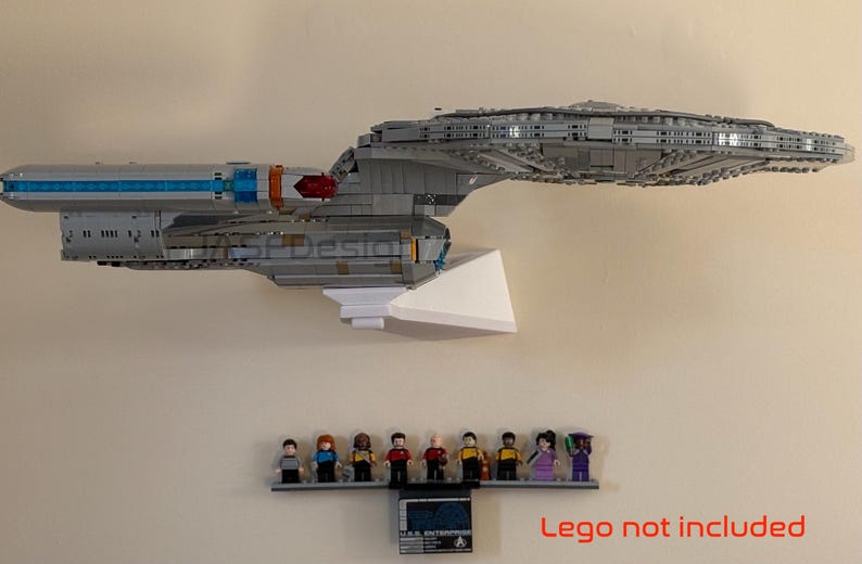 Puede incluir: Un modelo gris de una nave espacial, montado sobre un soporte blanco, con una peque&ntilde;a estanter&iacute;a debajo que contiene figuras en miniatura. El modelo tiene detalles en azul y naranja. La estanter&iacute;a tambi&eacute;n tiene una peque&ntilde;a placa. El texto "Lego not included" es visible.