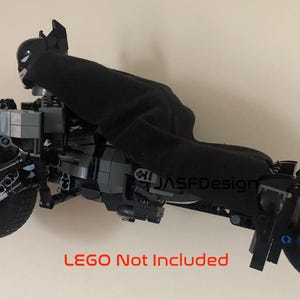 Op de afbeelding: Een zwart LEGO Batman minifiguur dat op een zwarte LEGO Batcycle rijdt. De Batcycle heeft een zwart en grijs ontwerp met een zwarte stoel en stuur. De Batcycle heeft twee wielen, &eacute;&eacute;n aan elke kant. De Batcycle staat op een witte achtergrond. LEGO niet inbegrepen.