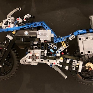 May include: A partially assembled LEGO motorcycle model with blue, black, and gray pieces. The motorcycle has a black front tire and a black rear tire. The motorcycle has a black seat and a black engine. The motorcycle has a gray luggage rack with a gray box on top. The box has the word "BMW" printed on it.