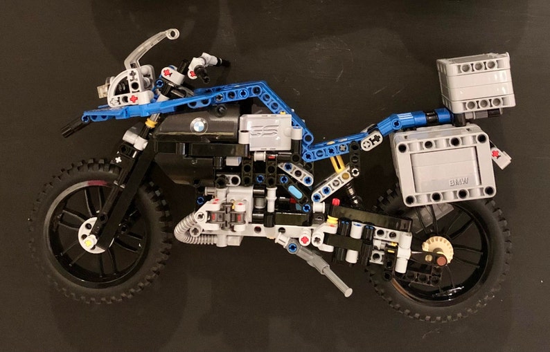 May include: A partially assembled LEGO motorcycle model with blue, black, and gray pieces. The motorcycle has a black front tire and a black rear tire. The motorcycle has a black seat and a black engine. The motorcycle has a gray luggage rack with a gray box on top. The box has the word "BMW" printed on it.