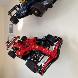 May include: Two Lego Formula One race cars, one red and one blue, are displayed on a white wall. The red car has the logos "V-Power", "HCLSoftware", and "Shell" on the side. The blue car has the logos "Oracle" and "Mobil" on the side.