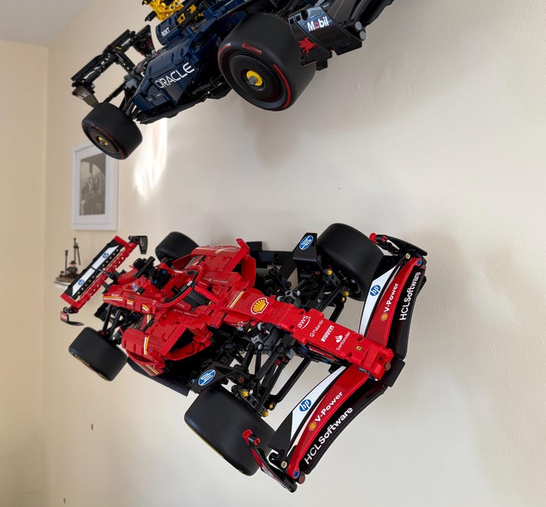 May include: Two Lego Formula One race cars, one red and one blue, are displayed on a white wall. The red car has the logos "V-Power", "HCLSoftware", and "Shell" on the side. The blue car has the logos "Oracle" and "Mobil" on the side.