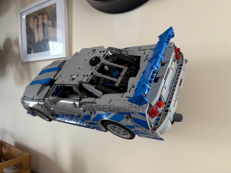 May include: A gray and blue model car made of interlocking plastic bricks. The car has a large blue spoiler and blue and white accents. The model is mounted on a wall. The text "JASFDesign" is visible on the side.