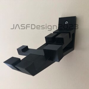 May include: A black 3D printed wall mount for a video game controller. The mount is attached to the wall with a screw. The mount is designed to hold the controller in a vertical position.