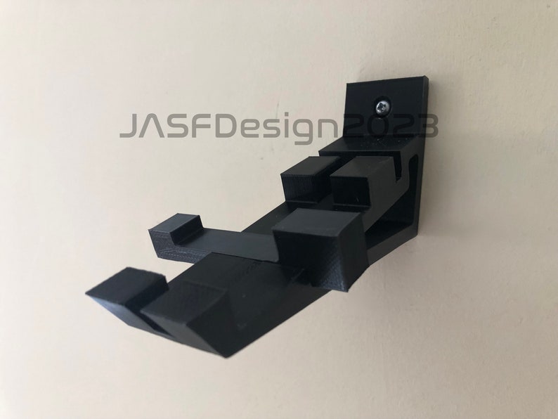 May include: A black 3D printed wall mount for a video game controller. The mount is attached to the wall with a screw. The mount is designed to hold the controller in a vertical position.