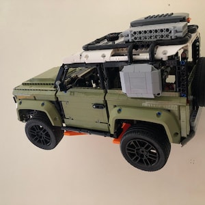 Puede incluir: Un detallado modelo Lego Technic de un veh&iacute;culo todoterreno verde oliva y blanco. El modelo presenta ruedas negras, una rueda de repuesto y una baca. El veh&iacute;culo est&aacute; suspendido contra una pared blanca.