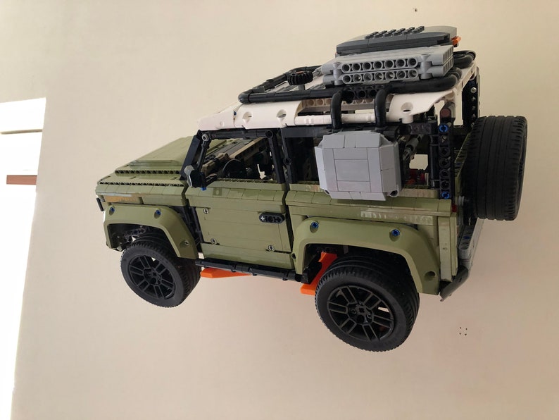 Puede incluir: Un detallado modelo Lego Technic de un veh&iacute;culo todoterreno verde oliva y blanco. El modelo presenta ruedas negras, una rueda de repuesto y una baca. El veh&iacute;culo est&aacute; suspendido contra una pared blanca.