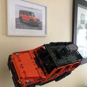 May include: A bright orange Lego model of a Mercedes-Benz G-Wagon, with black accents and a detailed interior, is mounted on a wall. The model is suspended in mid-air, giving the impression of flight.