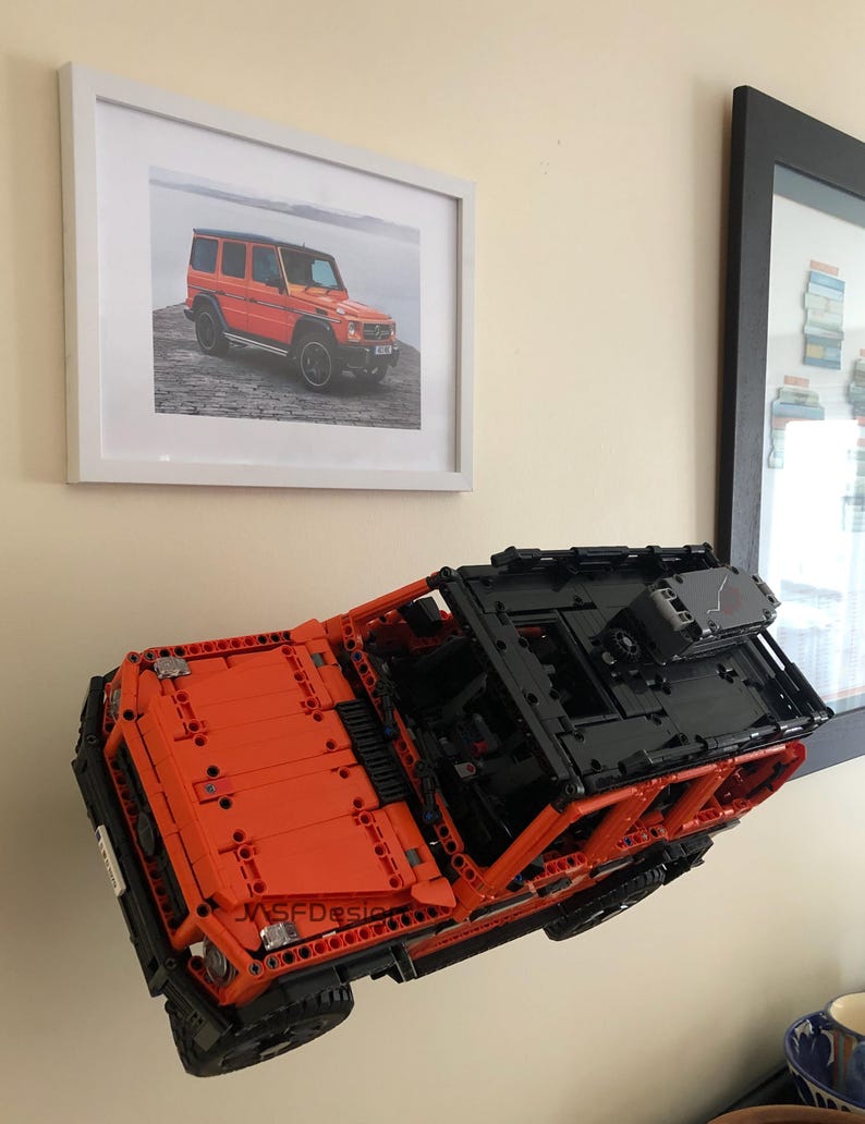 May include: A bright orange Lego model of a Mercedes-Benz G-Wagon, with black accents and a detailed interior, is mounted on a wall. The model is suspended in mid-air, giving the impression of flight.