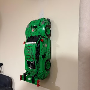 May include: A green Lego Porsche race car model mounted on a wall. The model features black wheels, red accents, and the word "PORSCHE" printed on the side. The car is displayed vertically.