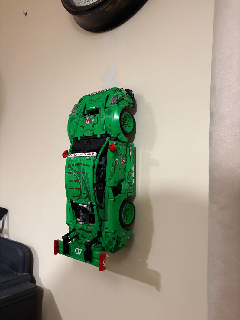 May include: A green Lego Porsche race car model mounted on a wall. The model features black wheels, red accents, and the word "PORSCHE" printed on the side. The car is displayed vertically.
