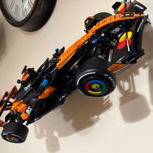 May include: A detailed model of a Formula 1 race car, predominantly orange and black, mounted on a wall. The car features sponsor logos, black tires, and a multi-colored wheel design. The model is angled, showcasing its aerodynamic design.