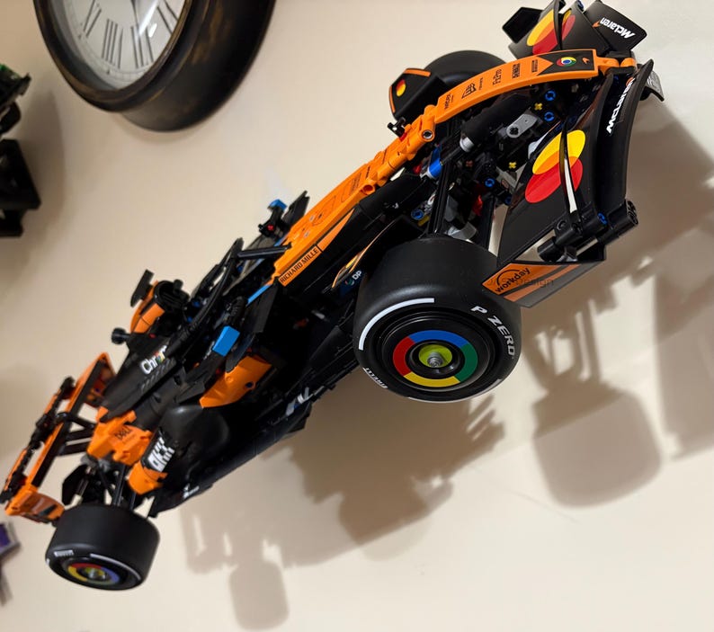 May include: A detailed model of a Formula 1 race car, predominantly orange and black, mounted on a wall. The car features sponsor logos, black tires, and a multi-colored wheel design. The model is angled, showcasing its aerodynamic design.