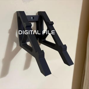 Op de afbeelding: Een zwarte wandbeugel met een geometrisch ontwerp. De beugel is met een enkele schroef aan een beige muur bevestigd. De tekst "DIGITAL FILE" is in het wit op de beugel gedrukt.