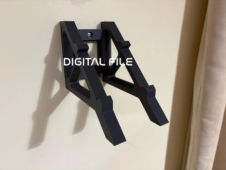 Op de afbeelding: Een zwarte wandbeugel met een geometrisch ontwerp. De beugel is met een enkele schroef aan een beige muur bevestigd. De tekst "DIGITAL FILE" is in het wit op de beugel gedrukt.