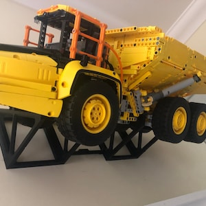 K&ouml;nnte beinhalten: Ein gelber Lego-Kipplaster mit schwarzem Fahrgestell und orangenen Akzenten wird auf einem schwarzen dreieckigen St&auml;nder pr&auml;sentiert. Der Laster ist leicht nach vorne geneigt und die Ladefl&auml;che ist angehoben.
