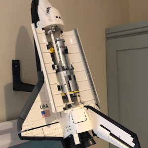 Puede incluir: Un modelo de transbordador espacial Lego blanco y negro con la palabra "USA" y una bandera estadounidense en el lateral. El transbordador est&aacute; montado en un soporte de pared negro.