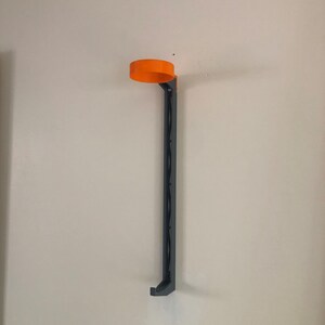 May include: A wall-mounted black metal holder with an orange plastic top. The holder is designed to hold a beverage can or bottle.