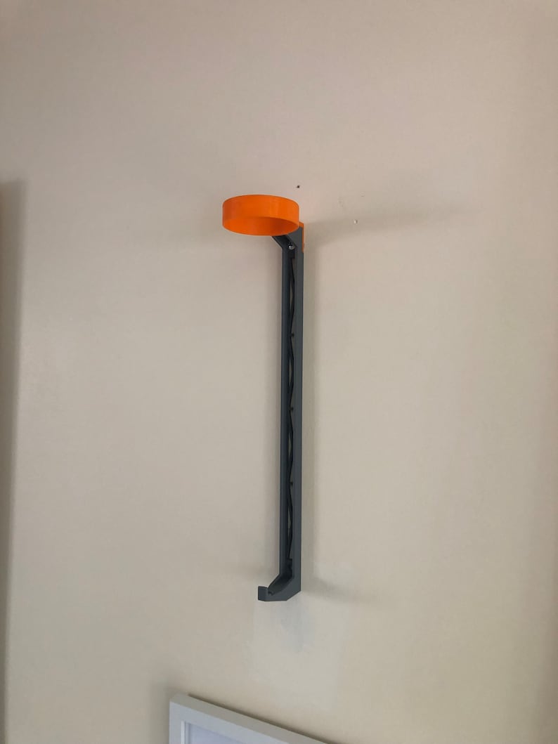 May include: A wall-mounted black metal holder with an orange plastic top. The holder is designed to hold a beverage can or bottle.