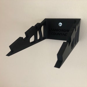 May include: Black wall mount for a Lamborghini Countach model car. The mount is made of plastic and has a cutout design. The text "Lamborghini Countach" is printed on the mount.