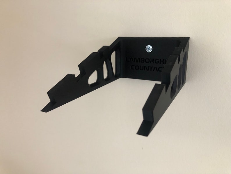 May include: Black wall mount for a Lamborghini Countach model car. The mount is made of plastic and has a cutout design. The text "Lamborghini Countach" is printed on the mount.