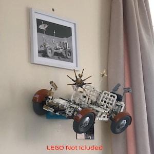 May include: A model of a lunar rover made of white and brown blocks, mounted on a wall. The rover has large wheels and various antennas. A framed black and white picture of a lunar rover is above it. The text "LEGO Not Included" is at the bottom.