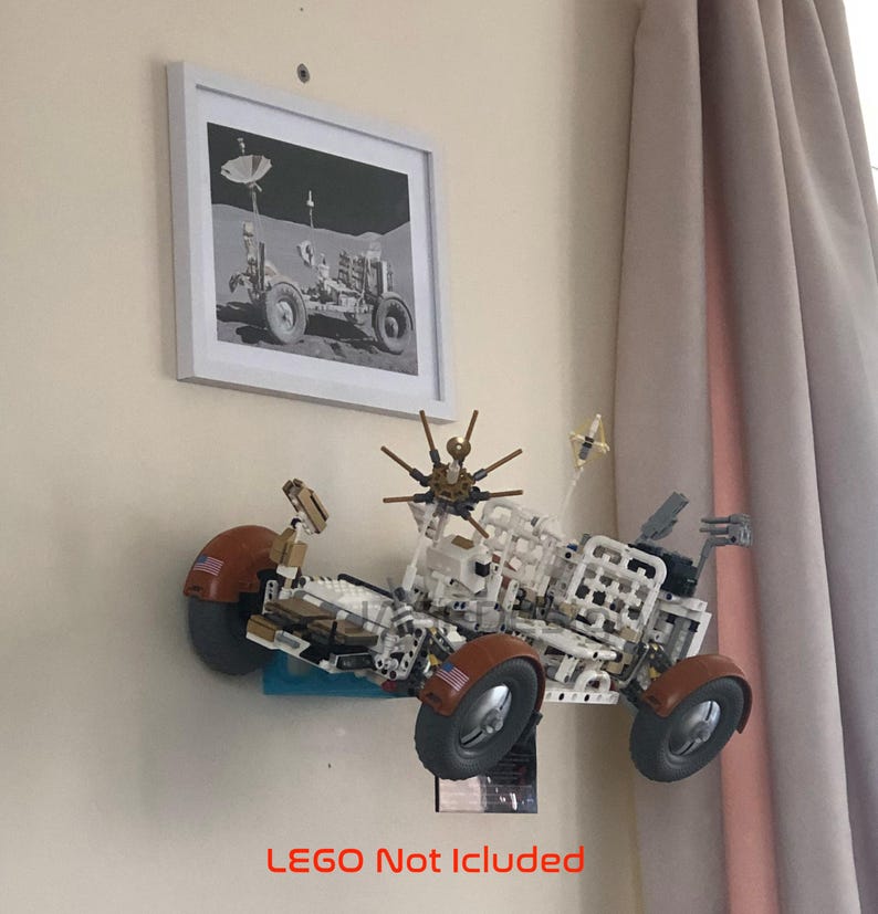 May include: A model of a lunar rover made of white and brown blocks, mounted on a wall. The rover has large wheels and various antennas. A framed black and white picture of a lunar rover is above it. The text "LEGO Not Included" is at the bottom.