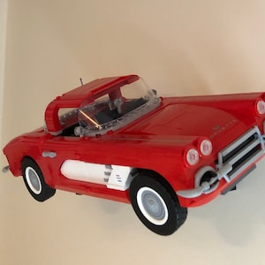 May include: A red Lego model of a classic Corvette convertible. The car has a white stripe and a clear windshield. The model is mounted on a wall.