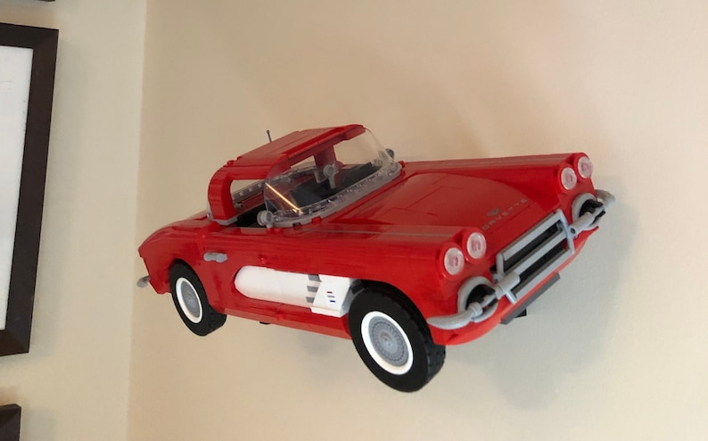 May include: A red Lego model of a classic Corvette convertible. The car has a white stripe and a clear windshield. The model is mounted on a wall.