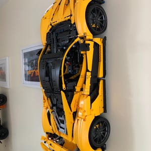May include: A large, yellow Lego model of a sports car mounted on a wall. The car features black wheels and intricate detailing, showcasing the underside of the vehicle. The model is displayed against a neutral wall, with a framed picture visible.