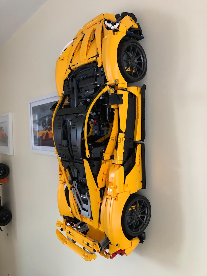 May include: A large, yellow Lego model of a sports car mounted on a wall. The car features black wheels and intricate detailing, showcasing the underside of the vehicle. The model is displayed against a neutral wall, with a framed picture visible.