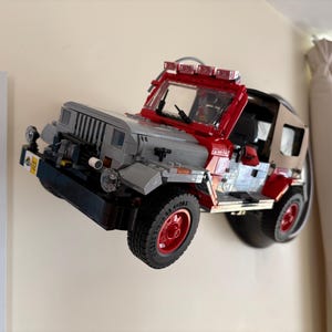 May include: A brick-built model of a Jurassic Park Jeep Wrangler mounted on a wall. The vehicle features a gray hood, red frame, tan soft top, and red wheels. A yellow license plate with the number 02 is visible on the front bumper.