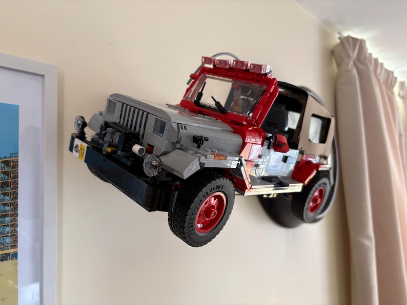 May include: A brick-built model of a Jurassic Park Jeep Wrangler mounted on a wall. The vehicle features a gray hood, red frame, tan soft top, and red wheels. A yellow license plate with the number 02 is visible on the front bumper.