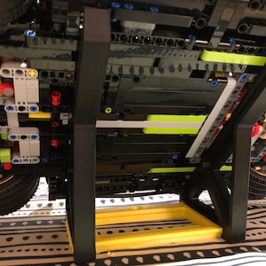 May include: A black Lego model car on a black display stand with a yellow base. The car is on a patterned fabric surface.