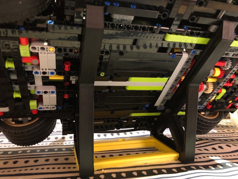 May include: A black Lego model car on a black display stand with a yellow base. The car is on a patterned fabric surface.