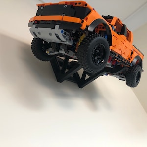 Op de afbeelding: Een oranje Lego Ford F-150 Raptor pick-up truck is gemonteerd op een zwarte driehoekige wandbeugel. De truck hangt in de lucht, met de voorkant naar de kijker.