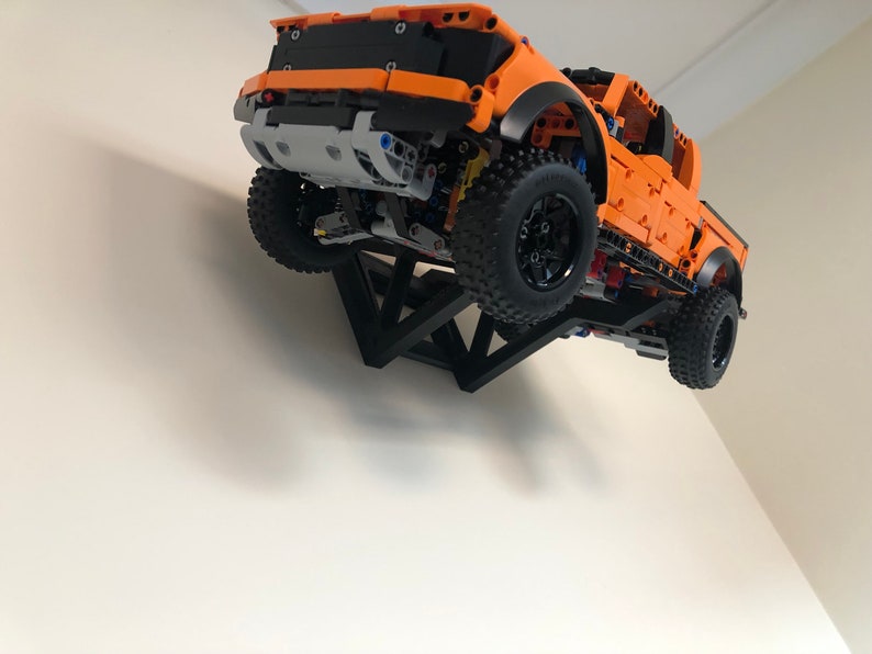 Op de afbeelding: Een oranje Lego Ford F-150 Raptor pick-up truck is gemonteerd op een zwarte driehoekige wandbeugel. De truck hangt in de lucht, met de voorkant naar de kijker.