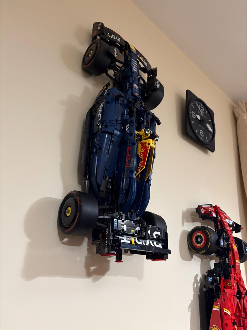 May include: Two Lego Formula 1 race cars mounted on a wall. The blue and yellow car is a Red Bull Racing car with the Oracle logo. The red car is a Ferrari car.