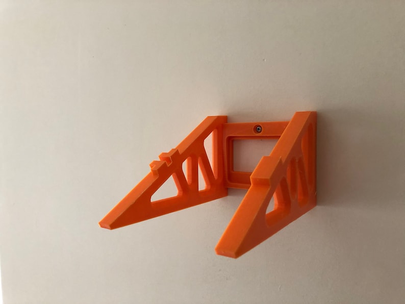 Op de afbeelding: Oranje 3D-geprint wandbevestiging voor een apparaat, zoals een telefoon of tablet. De beugel heeft een driehoekige vorm met open ruimtes en is met een schroef aan de muur bevestigd.