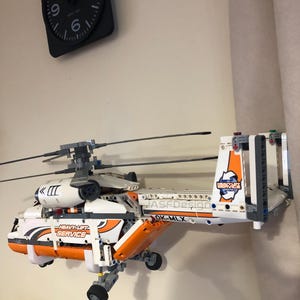 May include: A white and orange Lego helicopter model with the text "Heavy-Lift Service" and "OK-MLX" on the side. The helicopter is mounted on a wall and has a black and gray clock in the background.