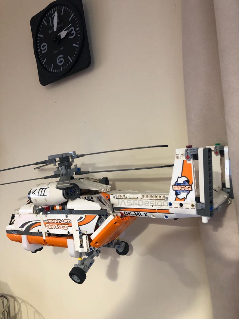 May include: A white and orange Lego helicopter model with the text "Heavy-Lift Service" and "OK-MLX" on the side. The helicopter is mounted on a wall and has a black and gray clock in the background.