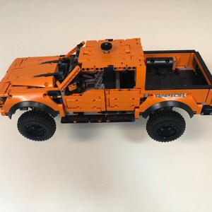 Op de afbeelding: Een oranje Lego Ford Raptor pick-up truck met zwarte wielen en een zwarte laadbak. De truck heeft een zwart "RAPTOR" logo aan de zijkant.