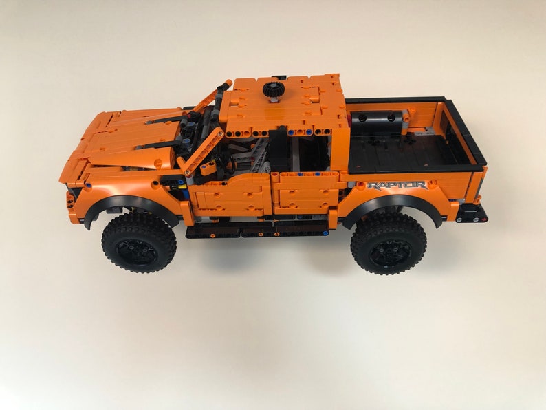 Op de afbeelding: Een oranje Lego Ford Raptor pick-up truck met zwarte wielen en een zwarte laadbak. De truck heeft een zwart "RAPTOR" logo aan de zijkant.
